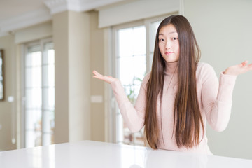 Beautiful Asian woman wearing casual sweater on white table clueless and confused expression with arms and hands raised. Doubt concept.