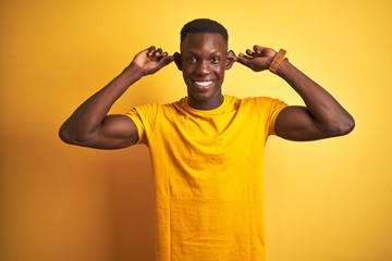 Fototapeta na wymiar Young african american man wearing casual t-shirt standing over isolated yellow background Smiling pulling ears with fingers, funny gesture. Audition problem