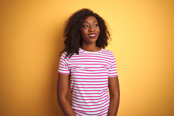 Young african american woman wearing striped t-shirt over isolated yellow background looking away to side with smile on face, natural expression. Laughing confident.