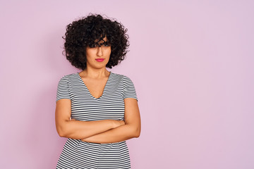 Young arab woman with curly hair wearing striped dress over isolated pink background skeptic and nervous, disapproving expression on face with crossed arms. Negative person.
