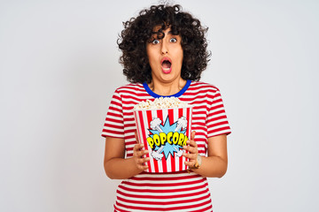 Young arab woman with curly hair holding pack of popcorns over isolated white background scared in shock with a surprise face, afraid and excited with fear expression