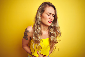 Young attactive woman wearing t-shirt standing over yellow isolated background with hand on stomach because indigestion, painful illness feeling unwell. Ache concept.