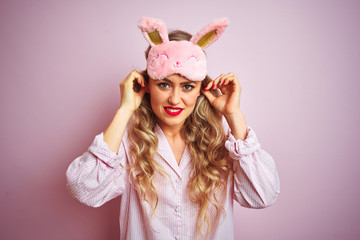 Young beautiful woman wearing pajama and sleep mask over pink isolated background covering ears with fingers with annoyed expression for the noise of loud music. Deaf concept.
