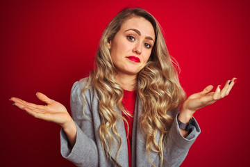 Young beautiful business woman wearing elegant jacket standing over red isolated background clueless and confused expression with arms and hands raised. Doubt concept.
