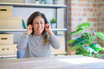 Middle age senior woman sitting at the table at home covering ears with fingers with annoyed expression for the noise of loud music. Deaf concept.