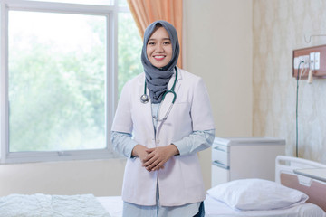 attractive young Asian Hijab female doctor smiling in front of camera inside hospital room with bed and windows as background