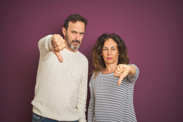 Beautiful middle age couple wearing winter sweater over isolated purple background looking unhappy and angry showing rejection and negative with thumbs down gesture. Bad expression.