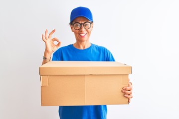 Senior deliverywoman wearing cap and glasses holding box over isolated white background doing ok sign with fingers, excellent symbol