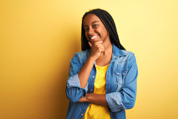Young african american woman wearing denim shirt standing over isolated yellow background looking confident at the camera smiling with crossed arms and hand raised on chin. Thinking positive.