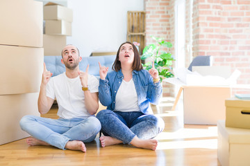 Young couple sitting on the floor arround cardboard boxes moving to a new house amazed and surprised looking up and pointing with fingers and raised arms.