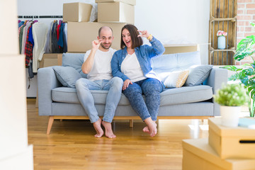 Young couple sitting on the sofa arround cardboard boxes moving to a new house smiling and confident gesturing with hand doing small size sign with fingers looking and the camera. Measure concept.