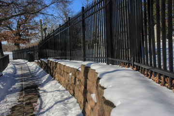 Snow covere walk way, bordered by a black rod iron fence with a blue sky and baren trees.