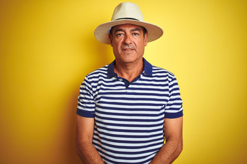 Handsome middle age man wearing striped polo and hat over isolated yellow background with serious expression on face. Simple and natural looking at the camera.