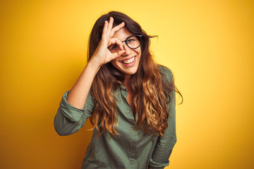 Young beautiful woman wearing green shirt and glasses over yelllow isolated background doing ok...