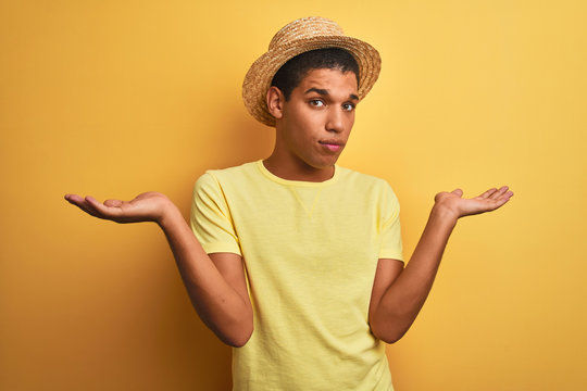 Young handsome arab man wearing t-shirt and summer hat over isolated yelllow background clueless and confused expression with arms and hands raised. Doubt concept.