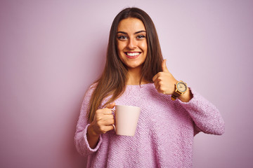 Young beautiful woman drinking cup of coffee standing over isolated pink background happy with big smile doing ok sign, thumb up with fingers, excellent sign