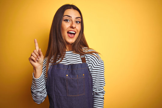 Young beautiful baker woman wearing apron standing over isolated yellow background pointing finger up with successful idea. Exited and happy. Number one.