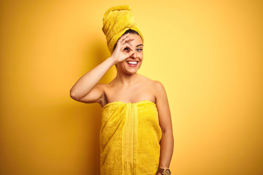 Young beautiful woman wearing towel after shower over isolated yellow background doing ok gesture with hand smiling, eye looking through fingers with happy face.