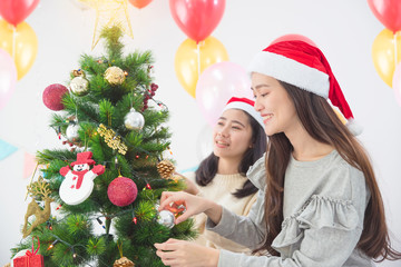 Beautiful asian girl with friend wearing Santa hats decorating Christmas tree at home for holidays.