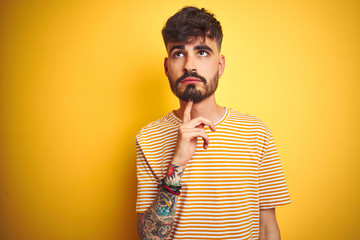 Young man with tattoo wearing striped t-shirt standing over isolated yellow background Thinking concentrated about doubt with finger on chin and looking up wondering