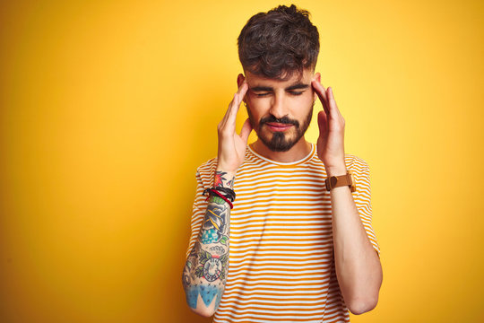 Young man with tattoo wearing striped t-shirt standing over isolated yellow background with hand on headache because stress. Suffering migraine.