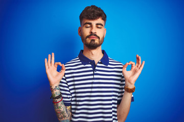 Fototapeta premium Young man with tattoo wearing striped polo standing over isolated blue background relax and smiling with eyes closed doing meditation gesture with fingers. Yoga concept.