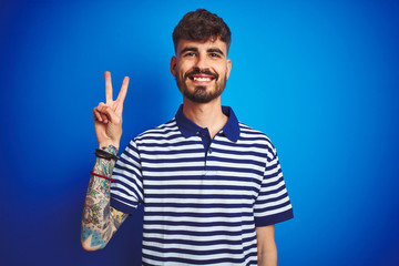 Young man with tattoo wearing striped polo standing over isolated blue background showing and pointing up with fingers number two while smiling confident and happy.