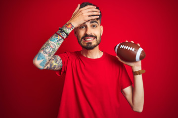 Young athlete man with tattoo holding football ball standing over isolated red background stressed with hand on head, shocked with shame and surprise face, angry and frustrated. Fear and upset 
