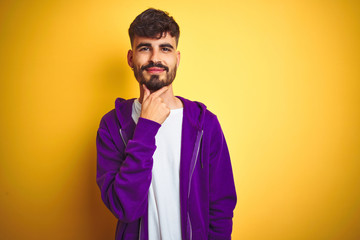 Young man with tattoo wearing sport purple sweatshirt over isolated yellow background looking confident at the camera with smile with crossed arms and hand raised on chin. Thinking positive.
