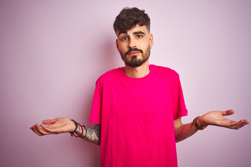 Young man with tattoo wearing t-shirt standing over isolated pink background clueless and confused...