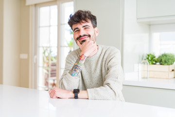 Young man wearing casual sweater sitting on white table looking confident at the camera with smile with crossed arms and hand raised on chin. Thinking positive.