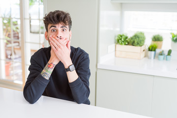 Young man wearing casual sweatshirt sitting on white table shocked covering mouth with hands for mistake. Secret concept.