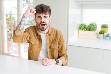Fototapeta premium Young man wearing casual jacket sitting on white table angry and mad raising fist frustrated and furious while shouting with anger. Rage and aggressive concept.