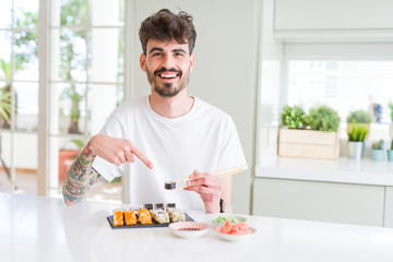Young man eating asian sushi from home delivery very happy pointing with hand and finger