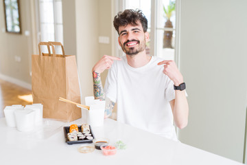 Young man eating asian sushi from home delivery looking confident with smile on face, pointing oneself with fingers proud and happy.