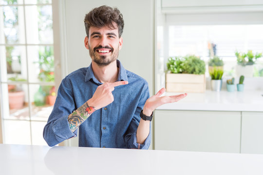 Young man wearing casual shirt sitting on white table amazed and smiling to the camera while presenting with hand and pointing with finger.