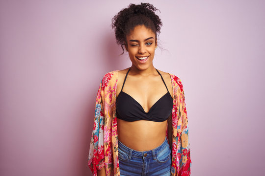 American woman on holiday wearing bikini and colorful dress over isolated pink background winking looking at the camera with sexy expression, cheerful and happy face.