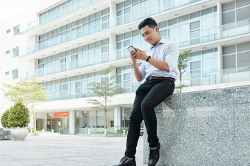 Smiling young Asian entrepreneur sitting outdoors and checking text messages in smartphone