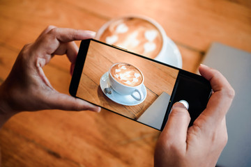 Hand taking a picture of hot latte art coffee cup with art foam on wooden table with her smart...