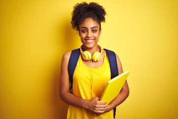 Afro woman using backpack and headphones holding notebook over isolated yellow background with a happy face standing and smiling with a confident smile showing teeth