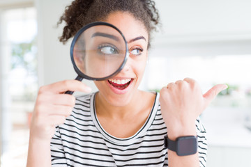 Young african american woman looking through magnifying glass pointing and showing with thumb up to...