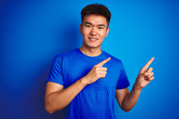 Young asian chinese man wearing t-shirt standing over isolated blue background smiling and looking at the camera pointing with two hands and fingers to the side.
