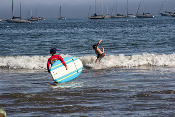 Swimmers heading into the ocean, 1 of them carrying a surfboard. White cap waves, and sailboats floating on the horizon.