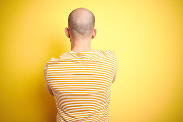 Young bald man with beard wearing casual striped t-shirt over yellow isolated background standing backwards looking away with crossed arms
