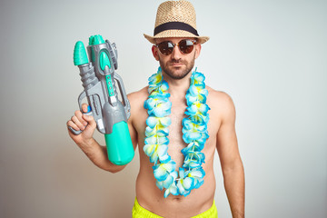 Young man wearing summer hat and hawaiian lei flowers holding water gun over isolated background...