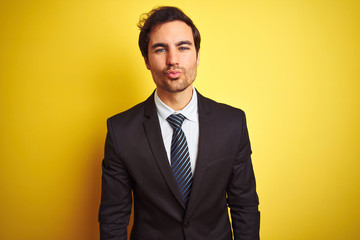 Young handsome businessman wearing suit and tie standing over isolated yellow background looking at the camera blowing a kiss on air being lovely and sexy. Love expression.