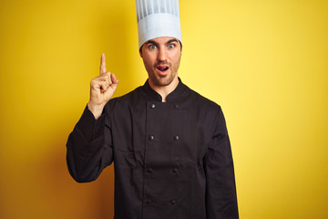 Young chef man wearing uniform and hat standing over isolated yellow background pointing finger up with successful idea. Exited and happy. Number one.