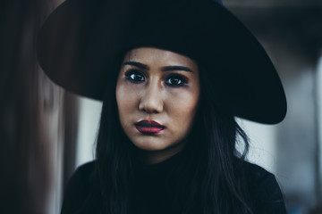 Long hair young woman in black dress and witch hat and red lips stand on aggressive face on black background, Halloween holiday. Horror theme.