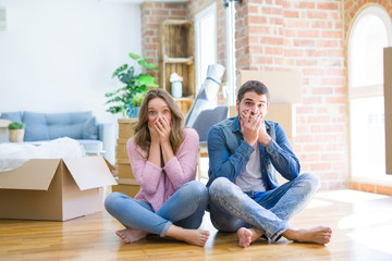 Young beautiful couple moving to a new house sitting on the floor shocked covering mouth with hands for mistake. Secret concept.