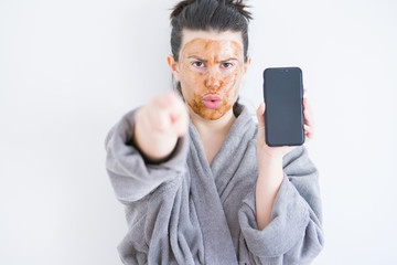 Beautiful woman wearing cosmetic facial mask as skincare treatment showing smartphone screen pointing with finger to the camera and to you, hand sign, positive and confident gesture from the front
